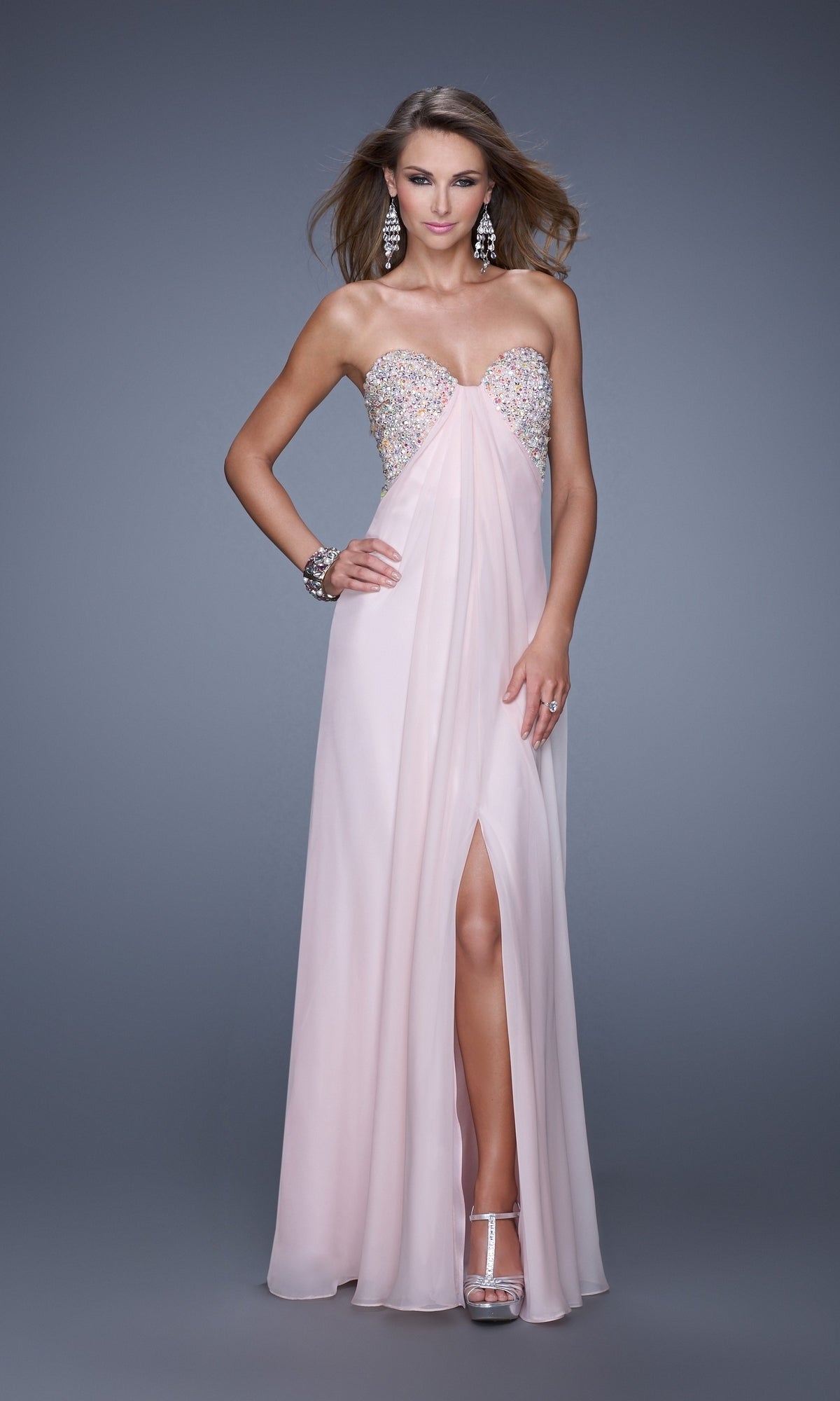 A woman in the La Femme Dress 20784, a strapless floor-length light pink chiffon gown with an embellished bust and front slit, pairs it with silver heels, statement earrings, and a bracelet against a gray background.